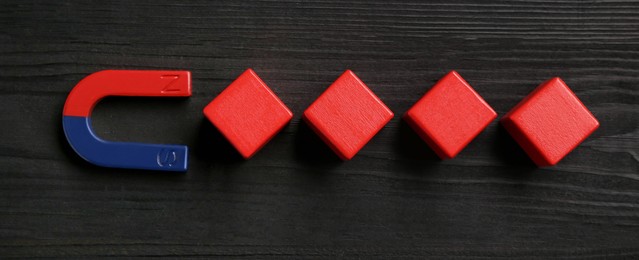 Photo of Magnet attracting red cubes on black wooden table, flat lay