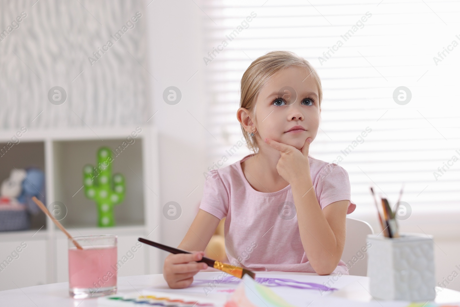 Photo of Cute girl thinking about her drawing at white table in kindergarten