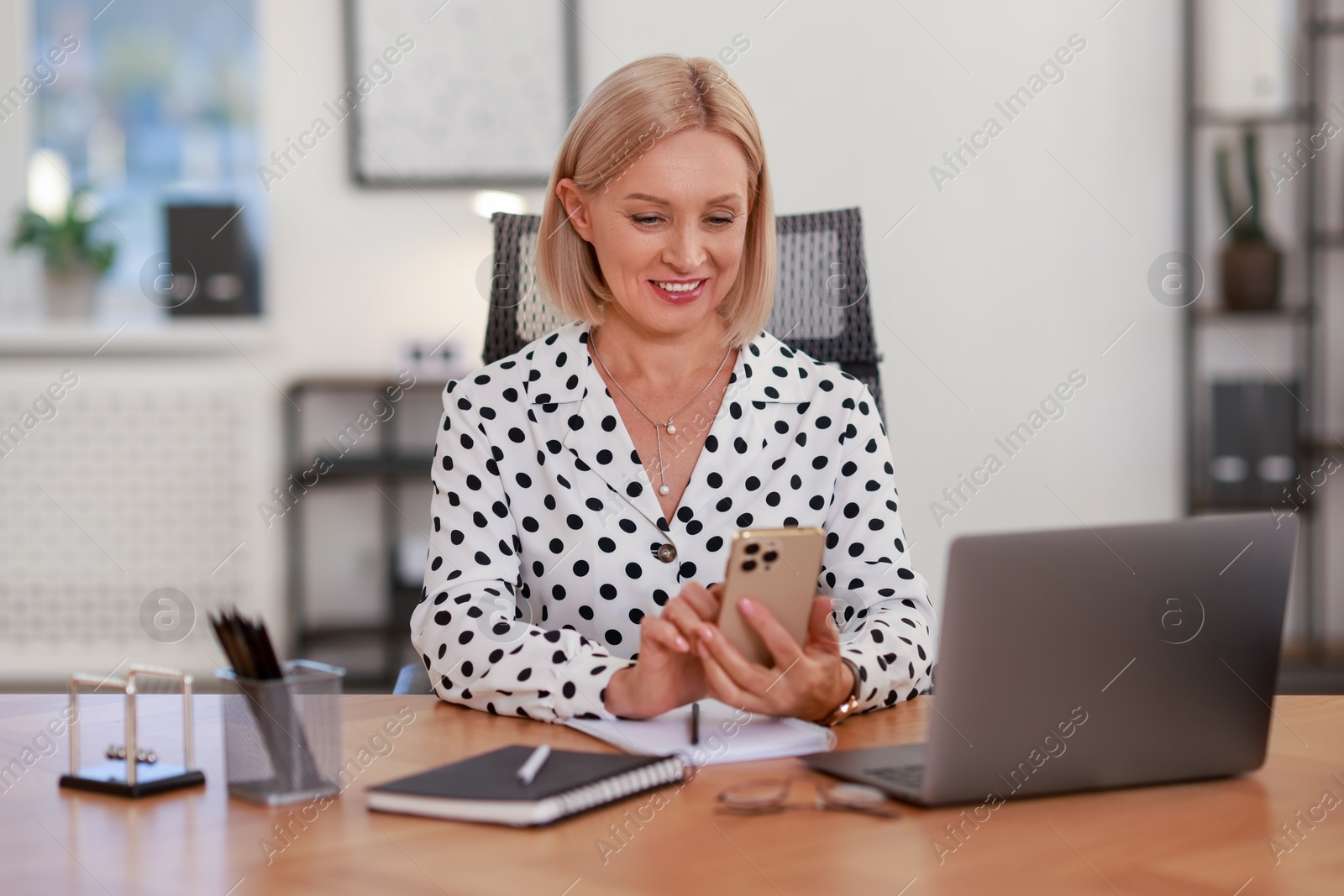 Photo of Smiling middle aged woman using smartphone at table in office