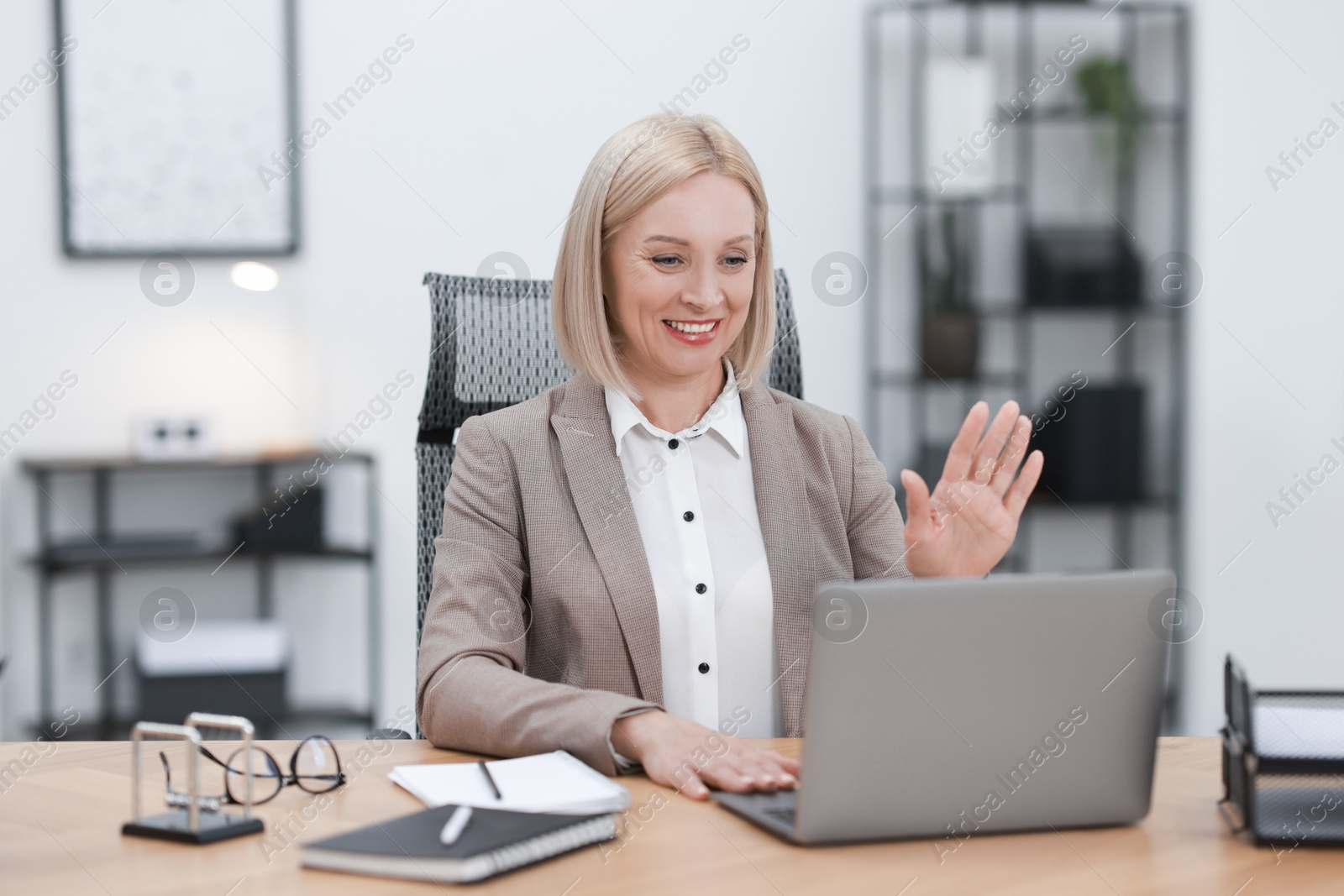 Photo of Smiling middle aged woman having videochat by laptop at table in office