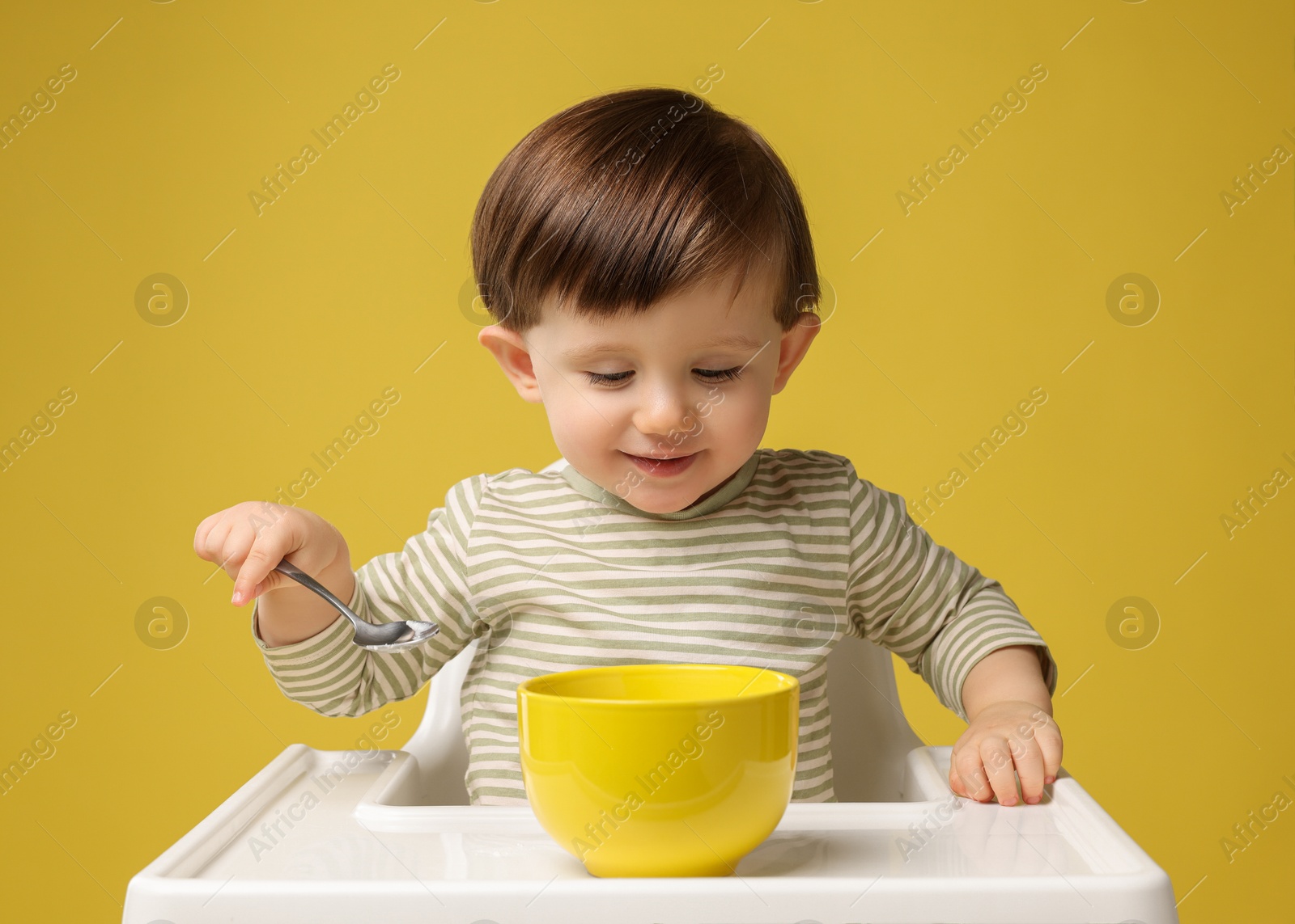 Photo of Cute little kid eating healthy baby food from bowl in high chair on yellow background