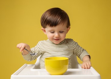 Cute little kid eating healthy baby food from bowl in high chair on yellow background