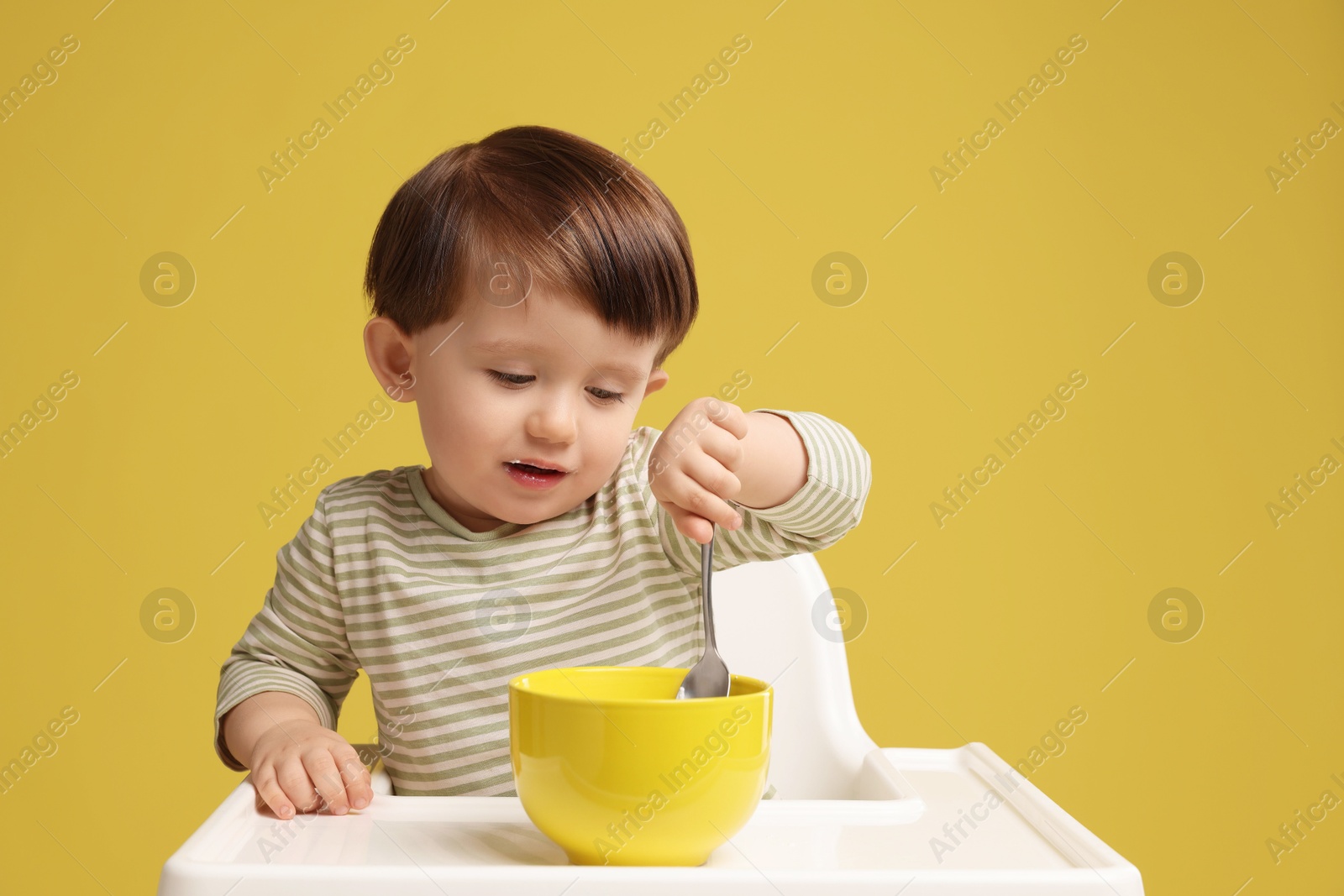 Photo of Cute little kid eating healthy baby food from bowl in high chair on yellow background