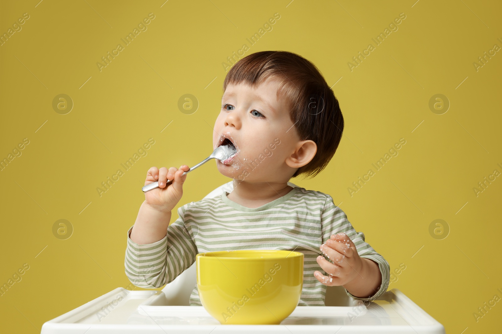 Photo of Cute little kid eating healthy baby food from bowl in high chair on yellow background