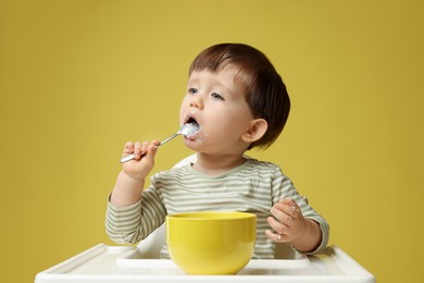 Photo of Cute little kid eating healthy baby food from bowl in high chair on yellow background