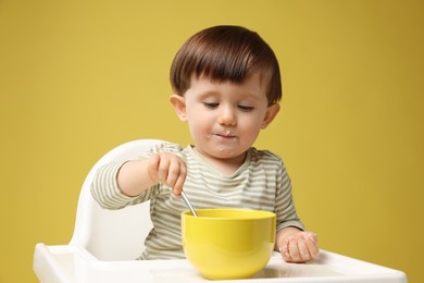 Photo of Cute little kid eating healthy baby food from bowl in high chair on yellow background