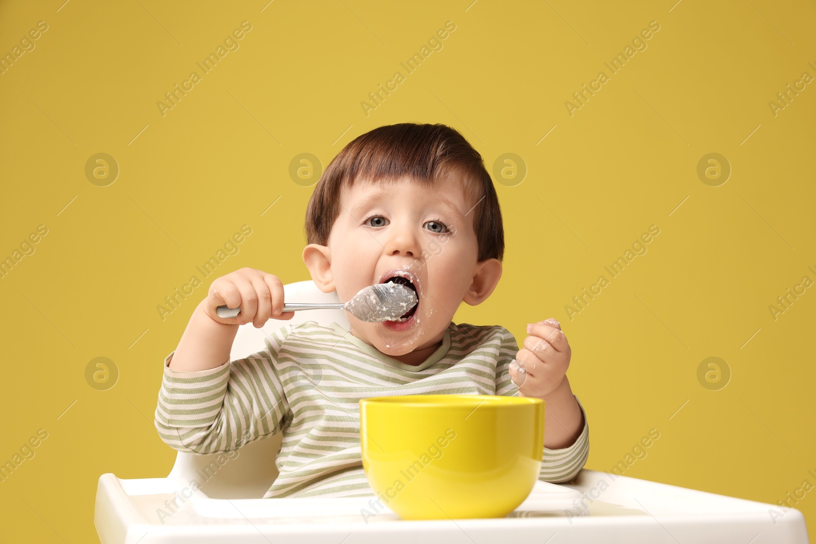 Photo of Cute little kid eating healthy baby food from bowl in high chair on yellow background