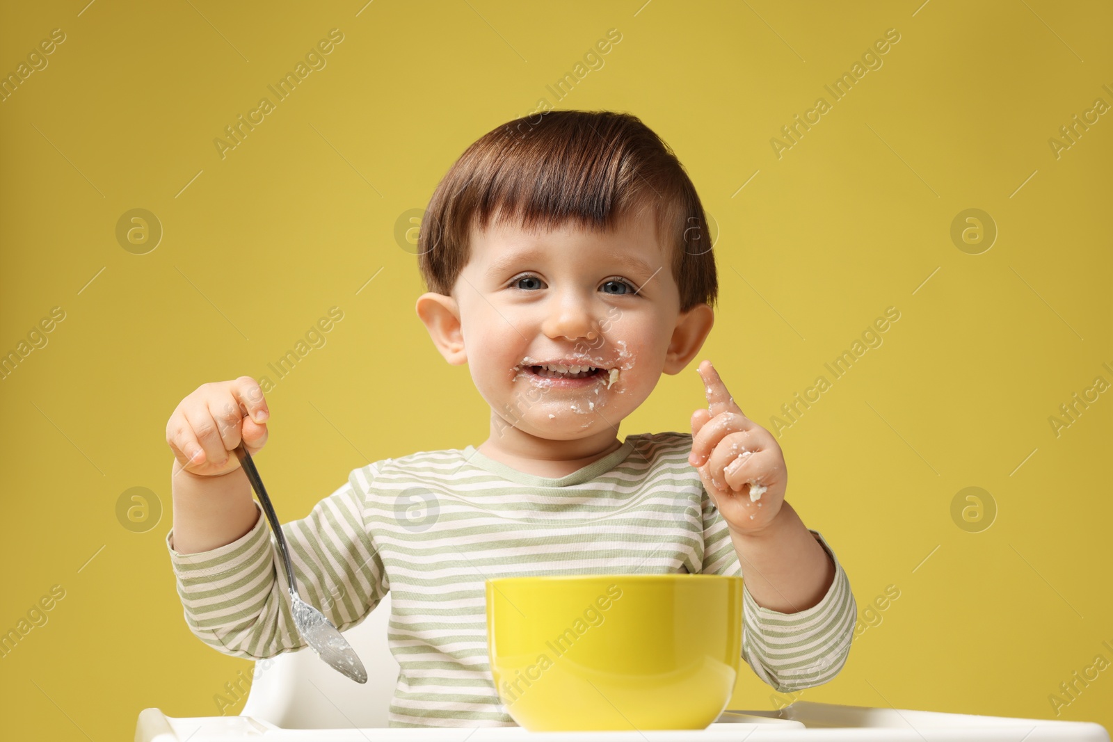 Photo of Cute little kid eating healthy baby food from bowl in high chair on yellow background