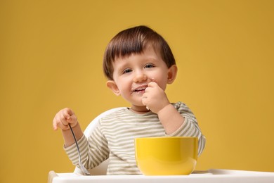 Photo of Cute little kid eating healthy baby food from bowl in high chair on yellow background