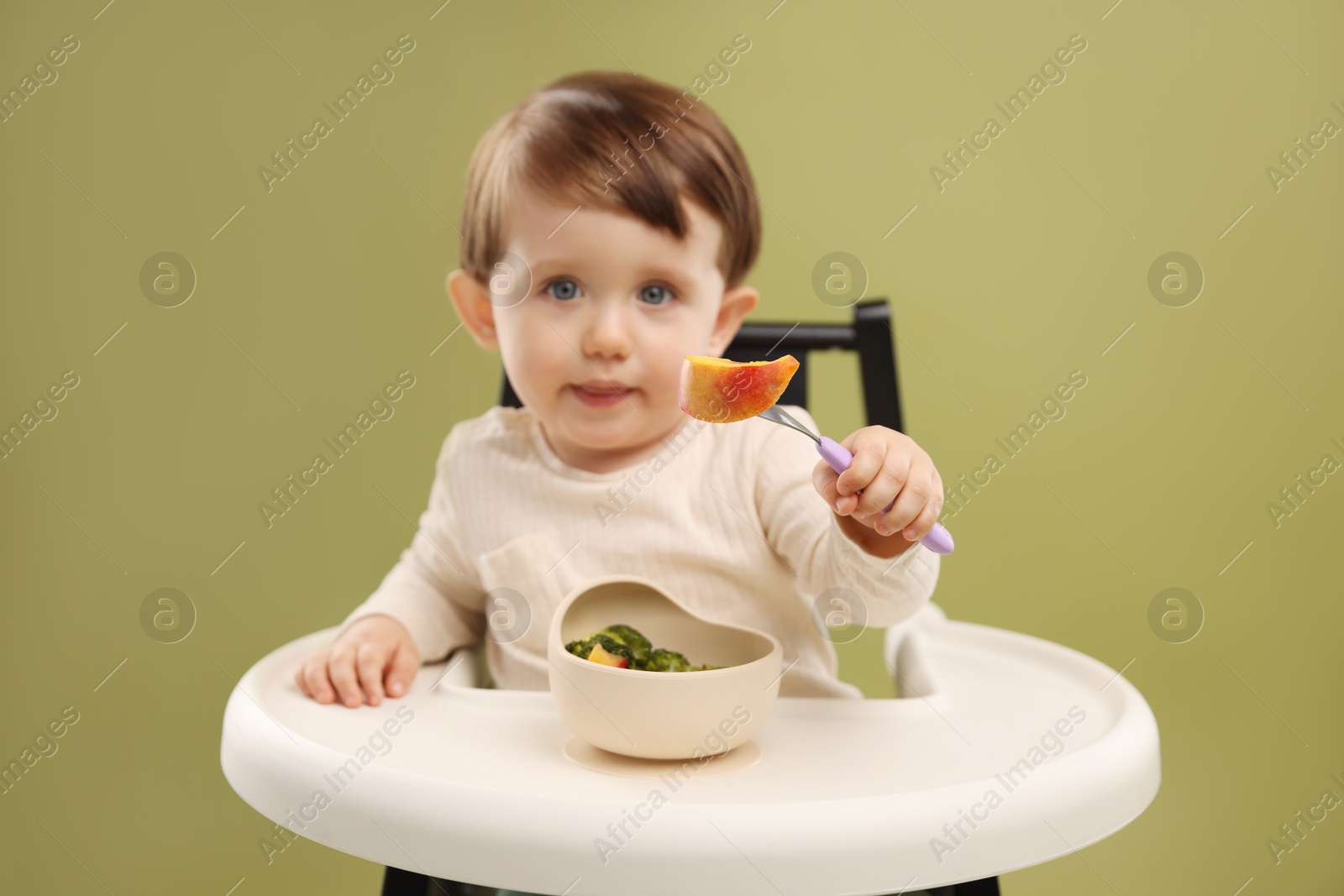 Photo of Cute little baby eating healthy food from bowl in high chair on olive background