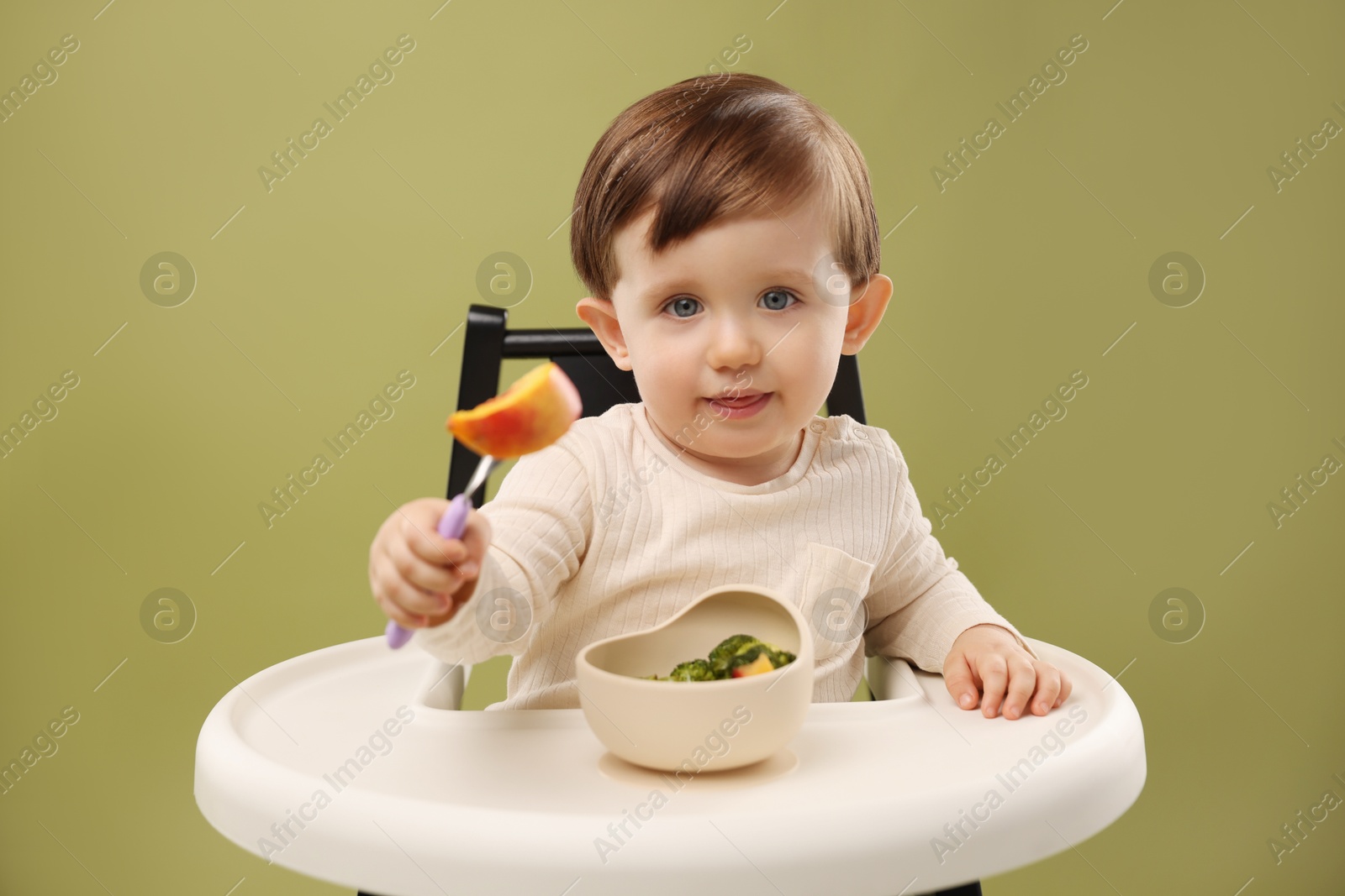 Photo of Cute little baby eating healthy food from bowl in high chair on olive background