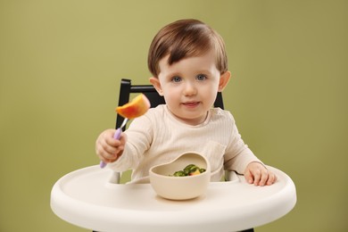 Photo of Cute little baby eating healthy food from bowl in high chair on olive background