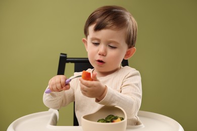 Photo of Cute little baby eating healthy food from bowl in high chair on olive background