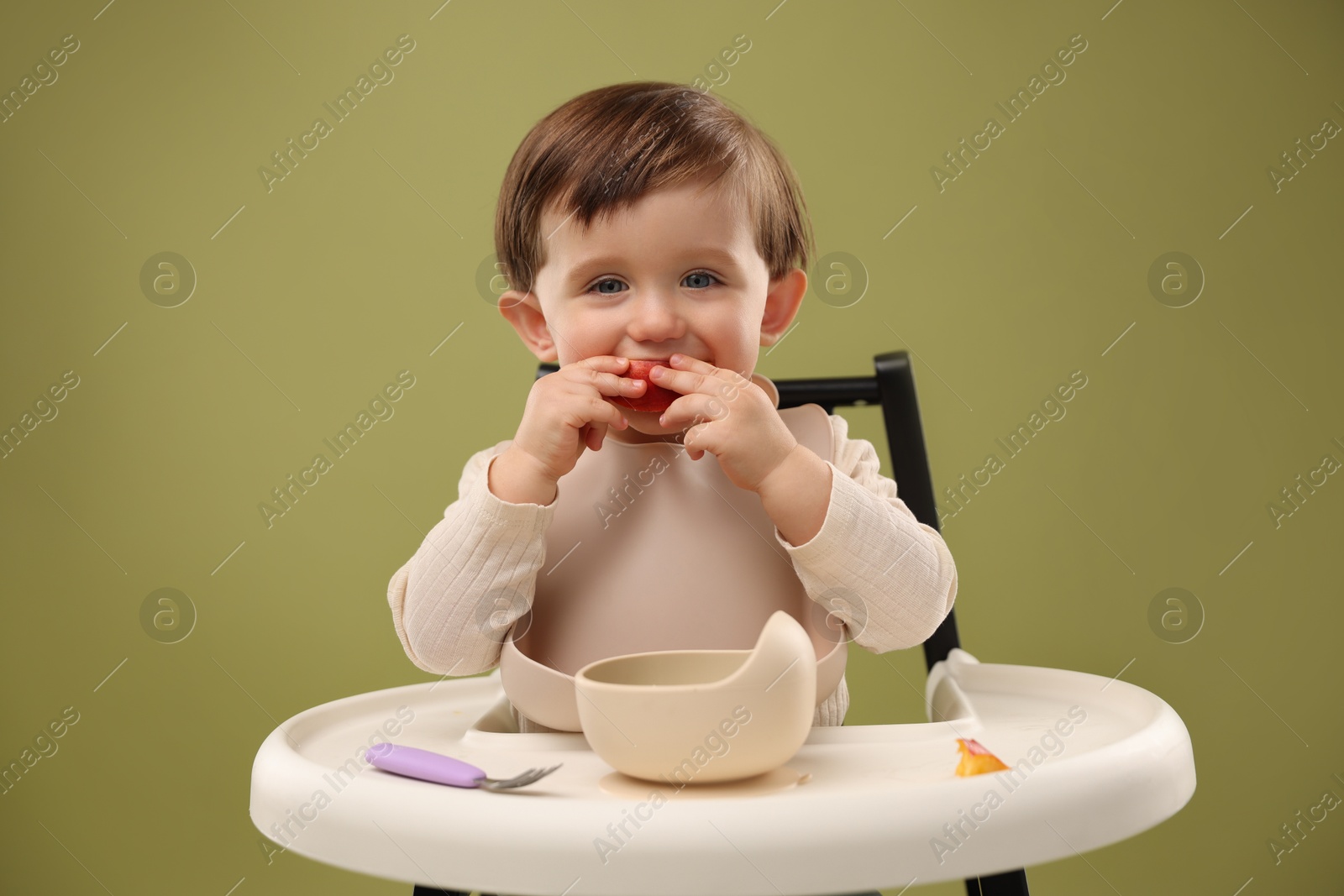 Photo of Cute little baby eating healthy food from bowl in high chair on olive background