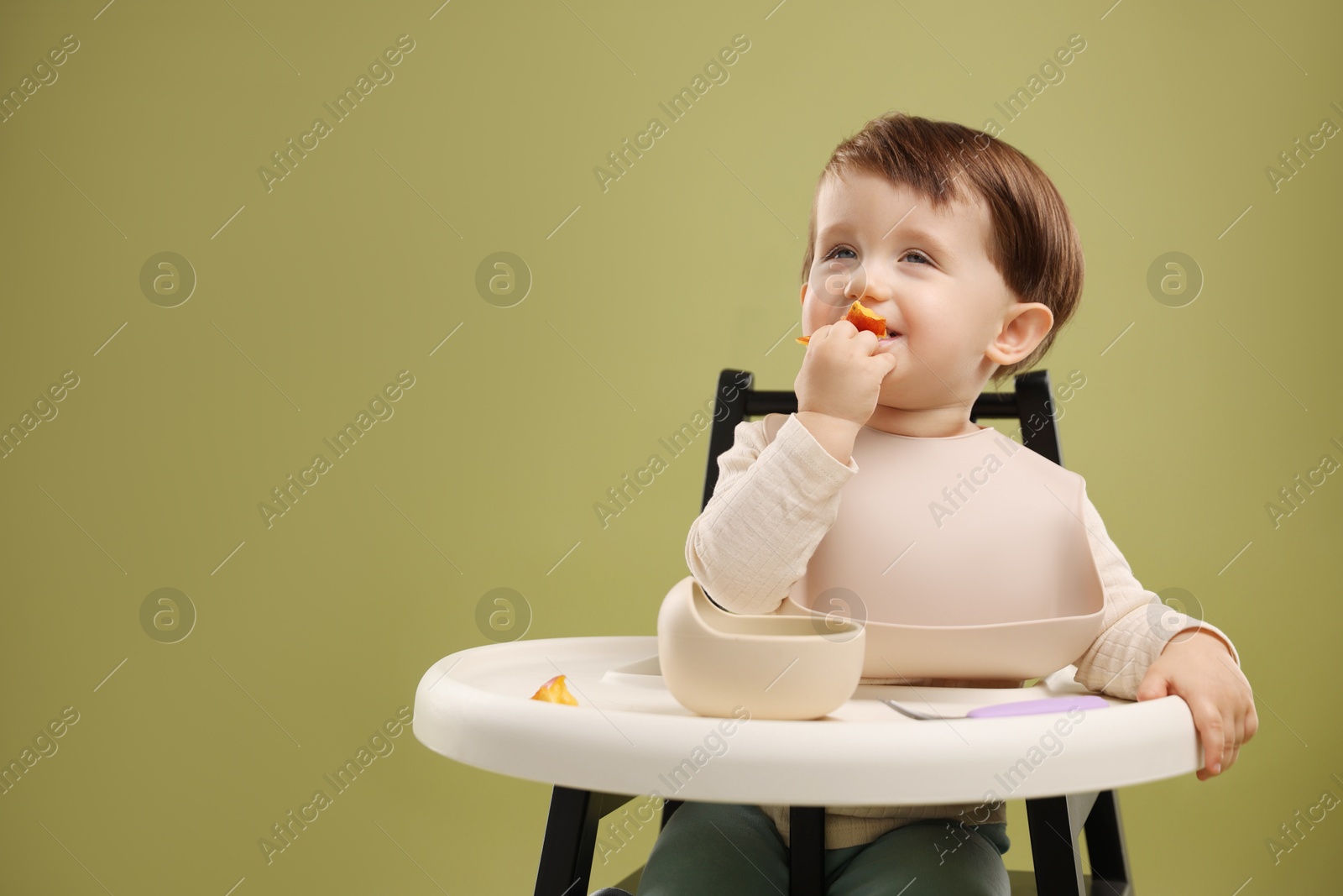 Photo of Cute little baby eating healthy food from bowl in high chair on olive background, space for text