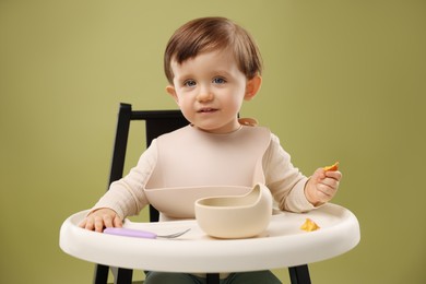 Photo of Cute little baby eating healthy food from bowl in high chair on olive background