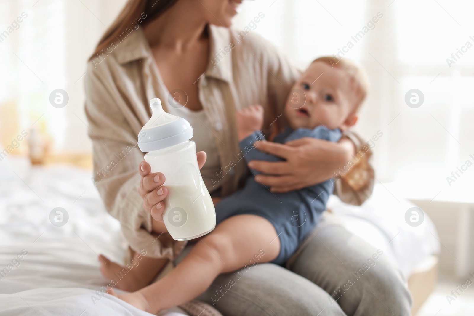 Photo of Mother holding cute little baby and bottle of milk on bed at home, selective focus