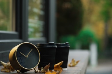 Photo of Autumn time. Alarm clock, fallen leaves and cups with hot drinks near cafe outdoors, space for text