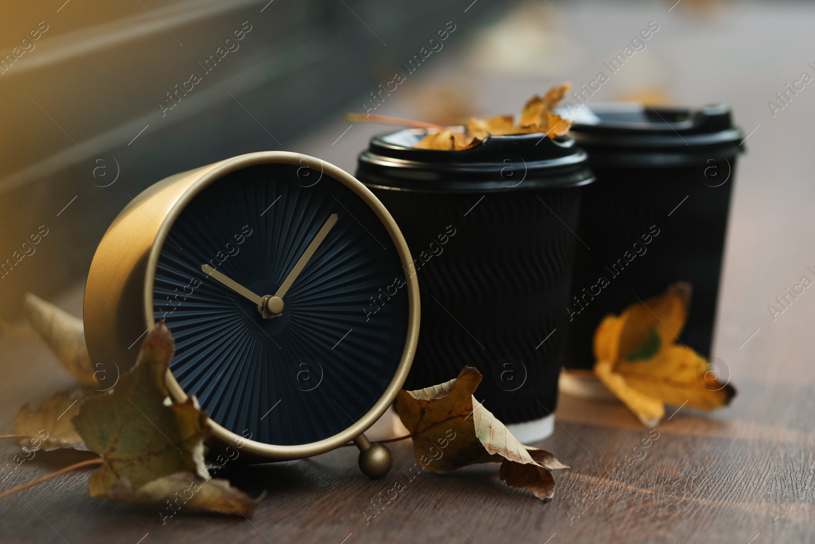 Photo of Autumn time. Alarm clock, fallen leaves and cups with hot drinks outdoors, space for text