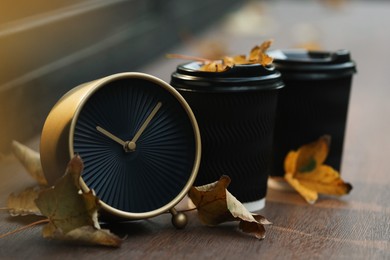 Photo of Autumn time. Alarm clock, fallen leaves and cups with hot drinks outdoors, space for text