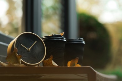 Photo of Autumn time. Alarm clock, fallen leaves and cups with hot drinks near cafe outdoors, space for text