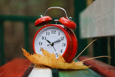 Photo of Autumn time. Alarm clock and fallen leaf on wooden bench outdoors