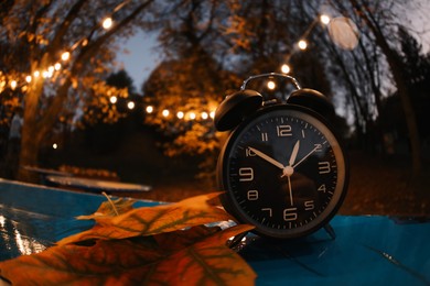 Photo of Autumn time. Alarm clock and fallen leaves on blue surface in evening park, wide angle lens