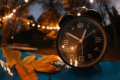 Photo of Autumn time. Alarm clock and fallen leaves on blue surface in evening park, wide angle lens