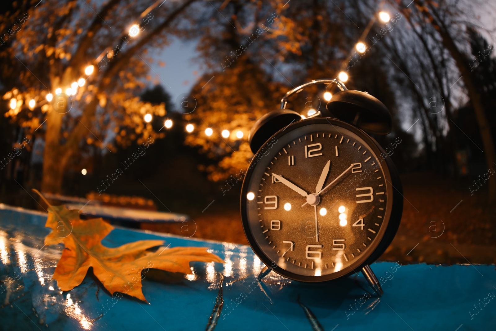 Photo of Autumn time. Alarm clock and fallen leaves on blue surface in evening park, wide angle lens