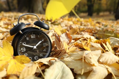 Photo of Alarm clock and yellow umbrella on dry leaves in park, closeup. Space for text