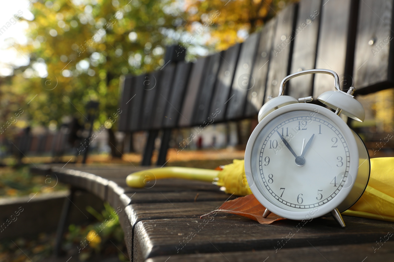 Photo of Alarm clock and yellow umbrella on wooden bench outdoors, closeup. Space for text