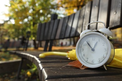 Photo of Alarm clock and yellow umbrella on wooden bench outdoors, closeup. Space for text