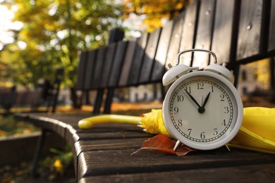 Photo of Alarm clock and yellow umbrella on wooden bench outdoors, closeup. Space for text