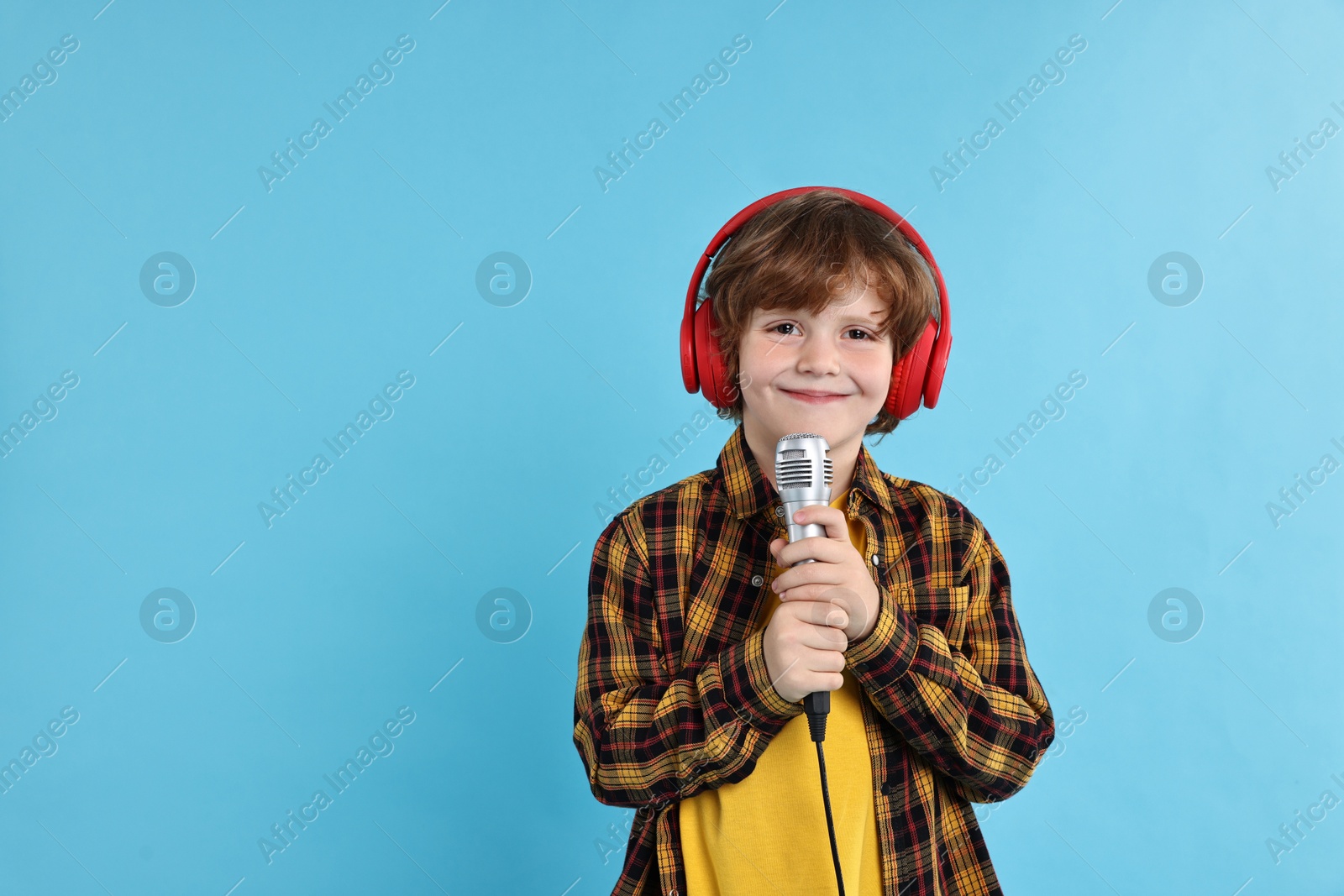 Photo of Little boy with microphone and headphones on light blue background, space for text