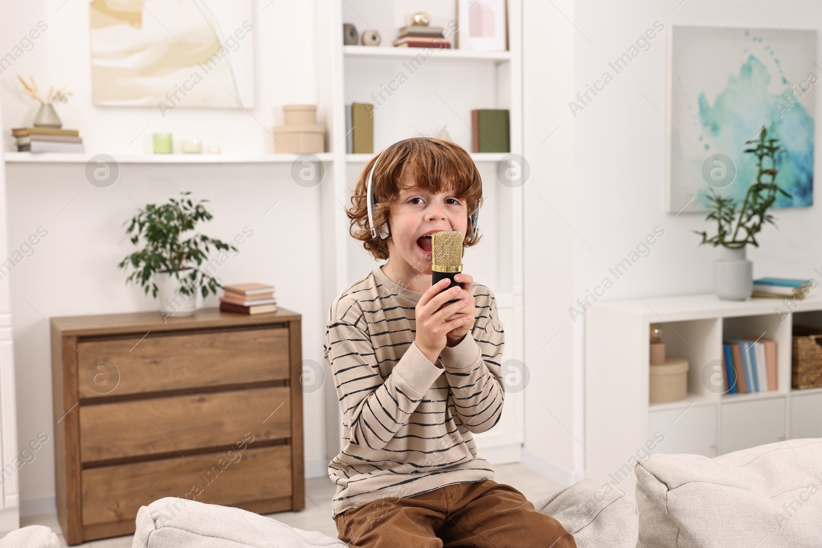 Photo of Little boy with microphone and headphones singing on sofa at home