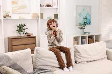 Photo of Little boy with microphone and headphones singing on sofa at home