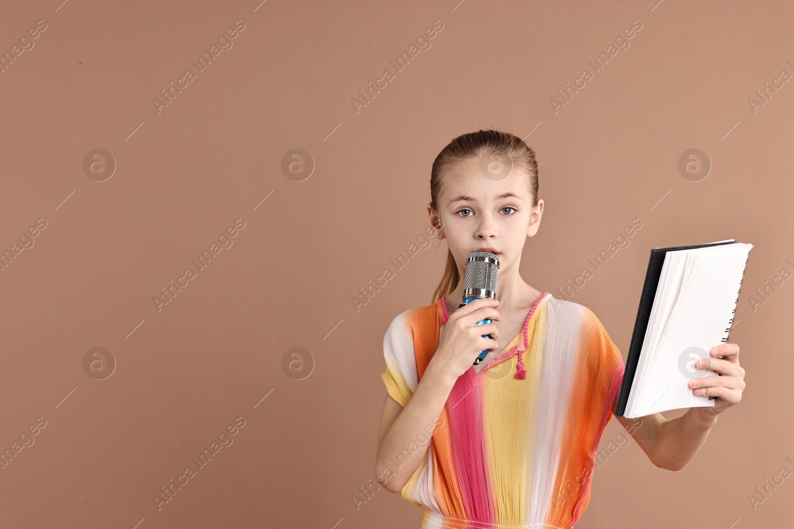 Photo of Little girl with microphone and notebook on light brown background, space for text