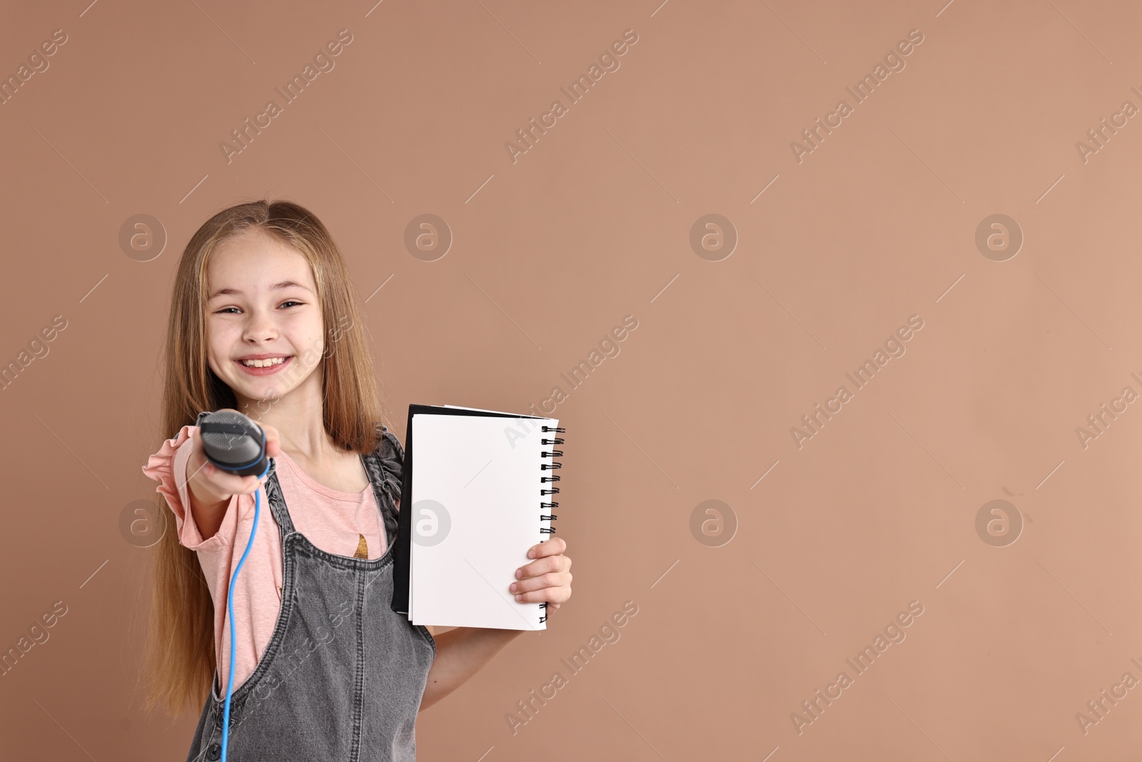 Photo of Little girl with microphone and notebook on light brown background, space for text