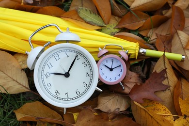 Photo of Alarm clocks and yellow umbrella on dry leaves in park, top view