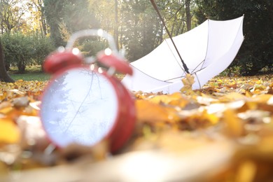 Photo of Alarm clock and white umbrella on dry leaves outdoors, selective focus. Space for text