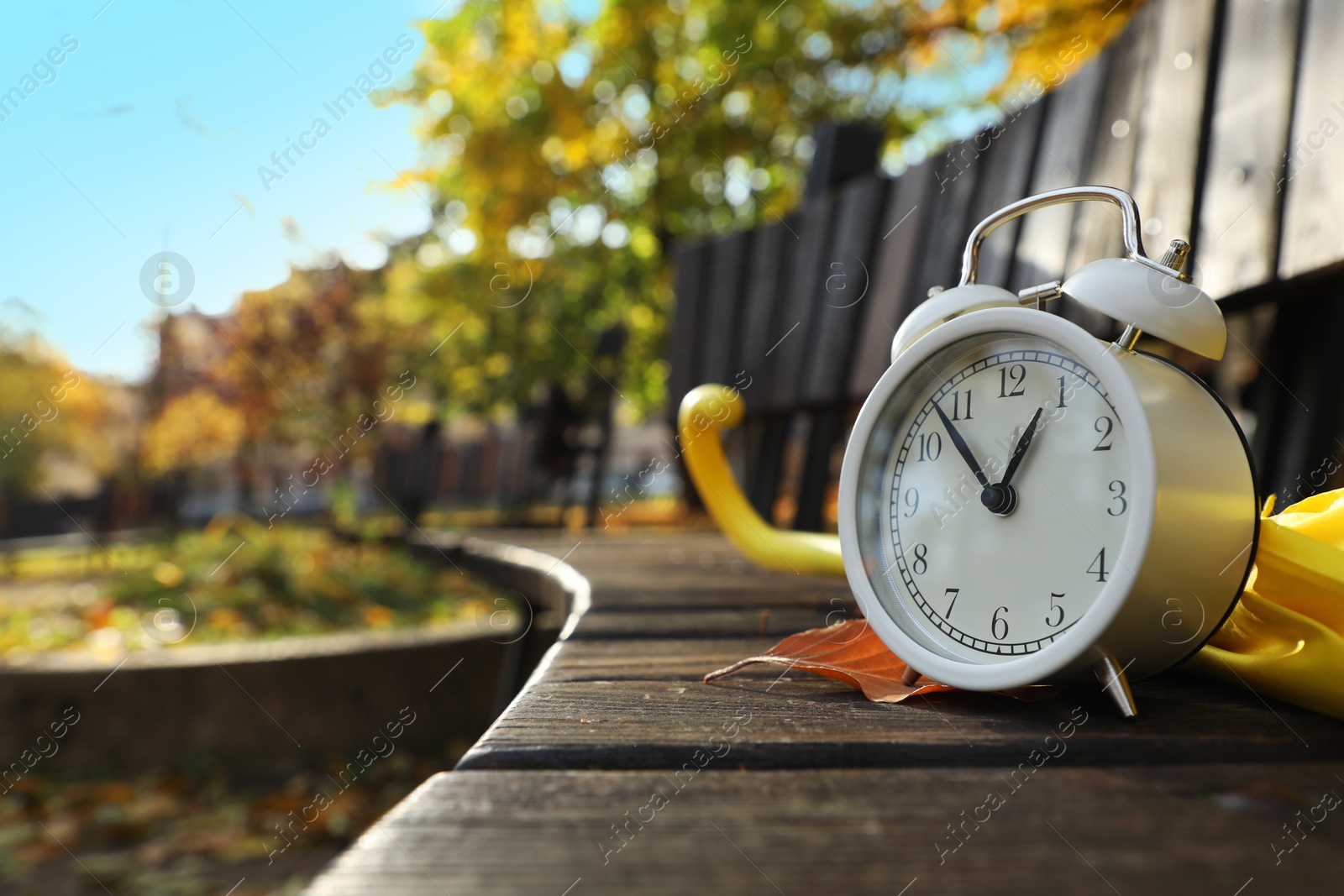 Photo of Alarm clock and yellow umbrella on wooden bench outdoors, closeup. Space for text