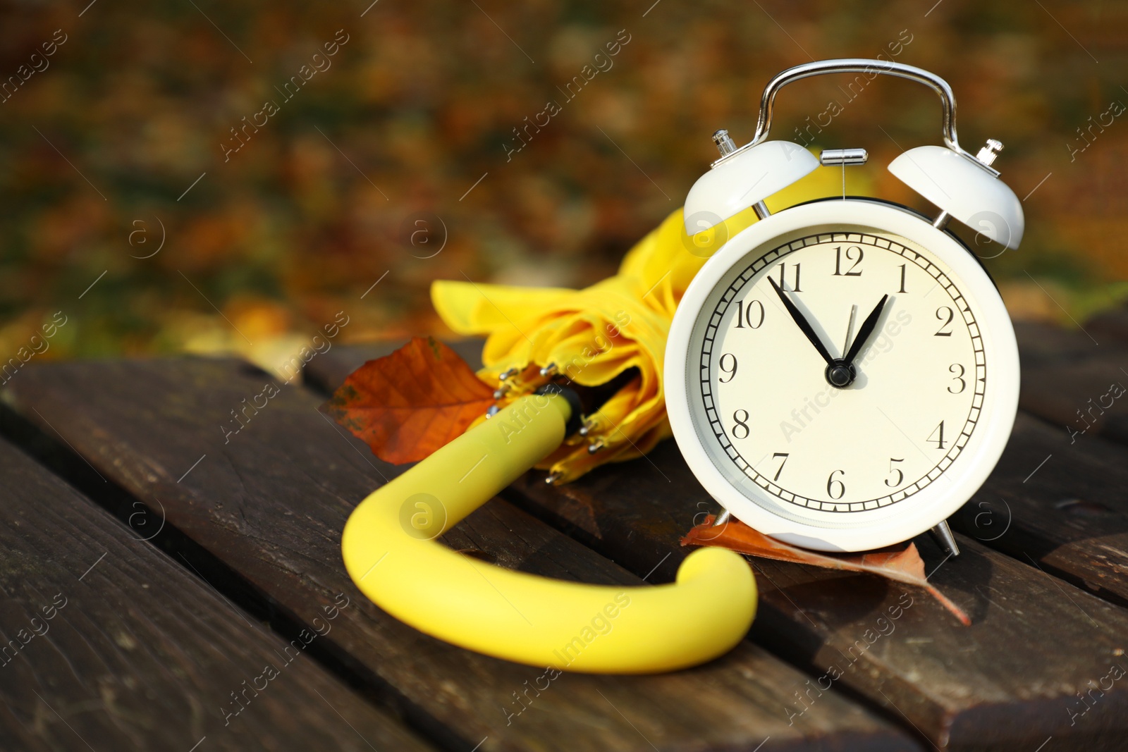 Photo of Alarm clock and yellow umbrella on wooden bench outdoors, closeup. Space for text