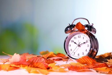 Photo of Alarm clock and dry leaves on white table against blurred background, closeup. Space for text