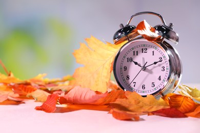 Photo of Alarm clock and dry leaves on white table against blurred background, closeup. Space for text