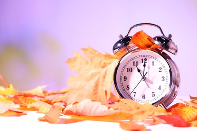 Photo of Alarm clock and dry leaves on white table against blurred background, closeup. Space for text