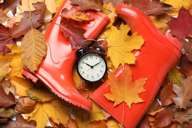 Photo of Alarm clock and rubber boots on dry leaves, flat lay