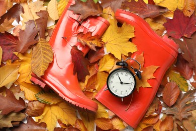 Photo of Alarm clock and rubber boots on dry leaves, flat lay