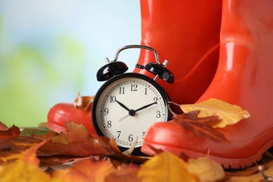 Photo of Alarm clock, dry leaves and rubber boots on blurred background, closeup