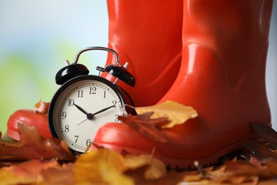 Photo of Alarm clock, dry leaves and rubber boots on blurred background, closeup