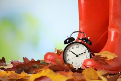 Photo of Alarm clock, dry leaves and rubber boots on blurred background, closeup. Space for text